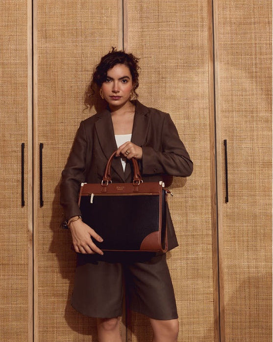 A woman stands in front of a woven wood cabinet, holding a structured black and brown leather briefcase-style handbag with top handles.
